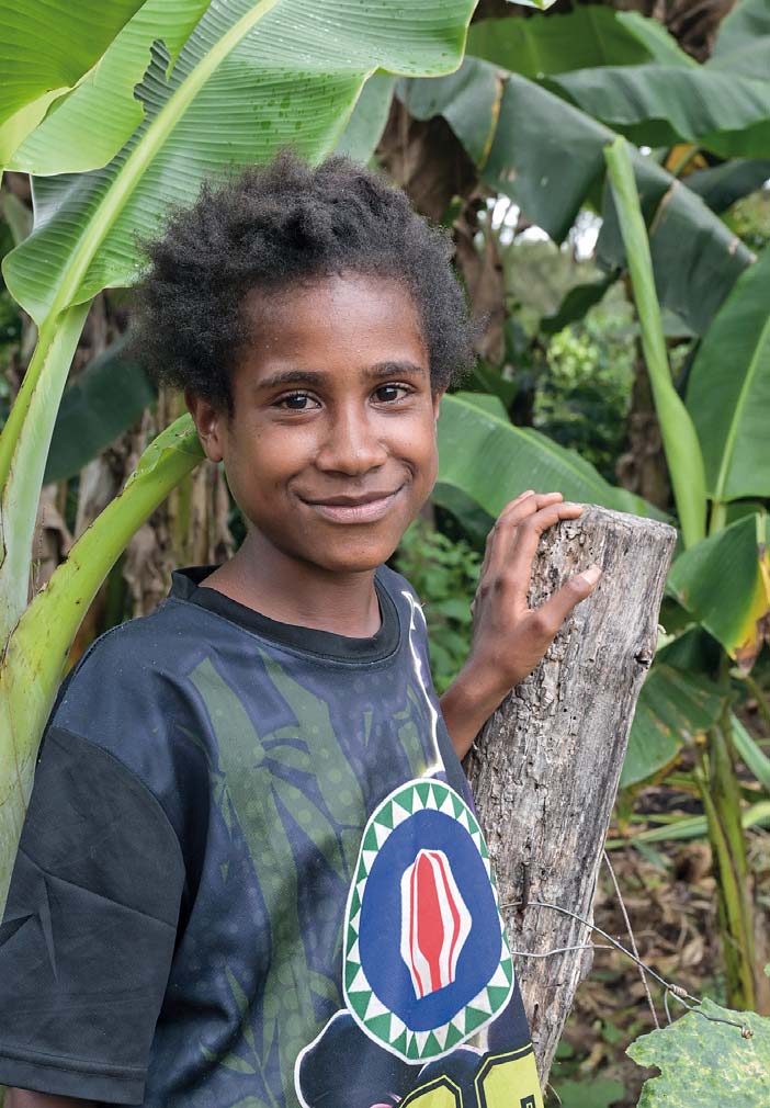 Ein Junge aus Papua steht an einen Zaunpfahl gelehnt und lächelt. Im Hintergrund Bananenstauden.
