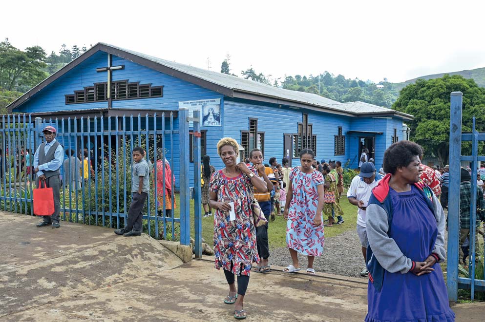 Menschen kommen aus einer Kirche, die durch ein blaues Tor von der Straße getrennt ist.