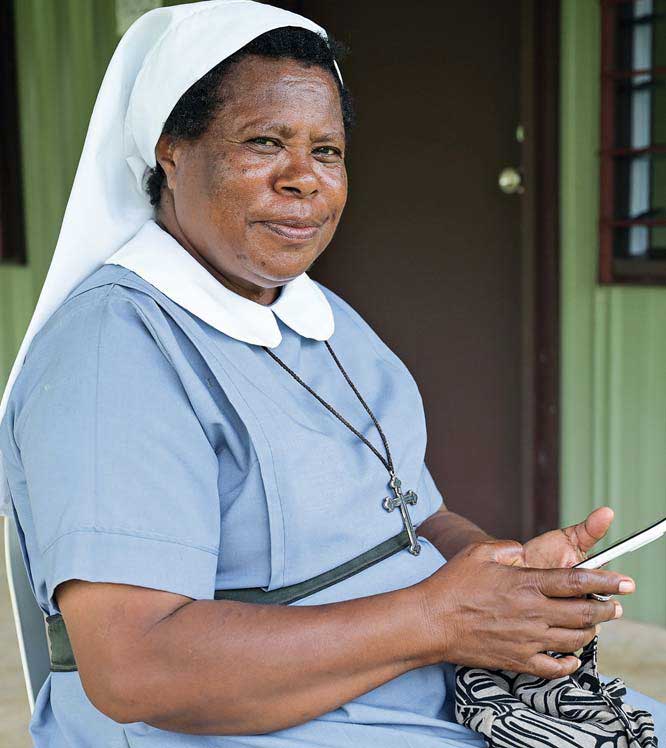 Schwester Thecla sitzt vor einem Gebäude. Sie trägt habit und blickt von ihrem Handy auf.