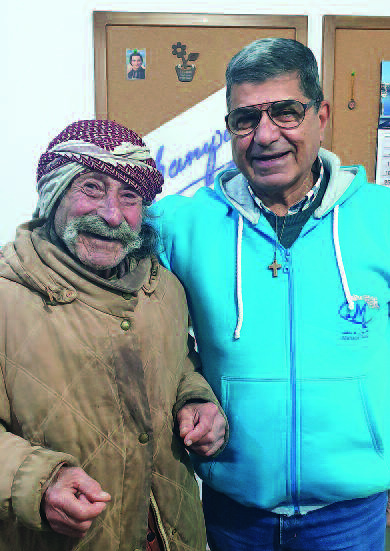 Links ein älterer Mann mit Schnauzbart, Kopftuch und schon länger getragener Jacke. Rechts von ihm ein Mann in mittlerem Alter mit Brille und einer Kette mit Holzkreuz um den Hals, der den Arm um ihn legt. Beide lächeln.