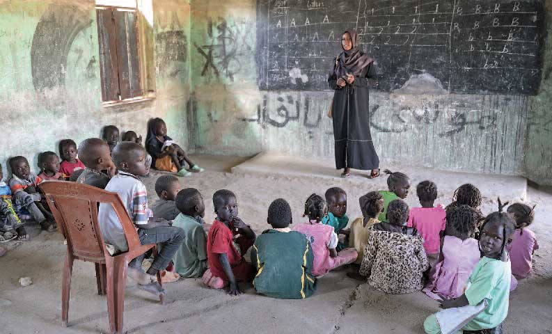 Blick in ein einfaches Klassenzimmer. Vor der Tafel steht die Lehrerin. Zwei Kinder teilen sich einen Plastikstuhl, alle übrigen sitzen auf dem Boden.