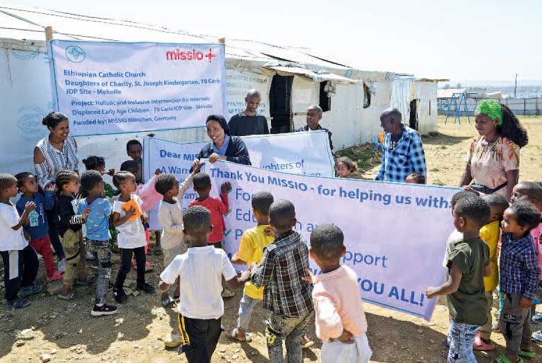 Projektpartnerinnen und -partner haben sich gemeinsam mit Kindern aus dem St. Josef Kindergarten vor Zelten versammelt, um Vertreter von missio zu begrüßen. Es sind große Transparente zu sehen mit der Aufschrift missio und Details um Projekt sowie ein Dank für die Hilfe.