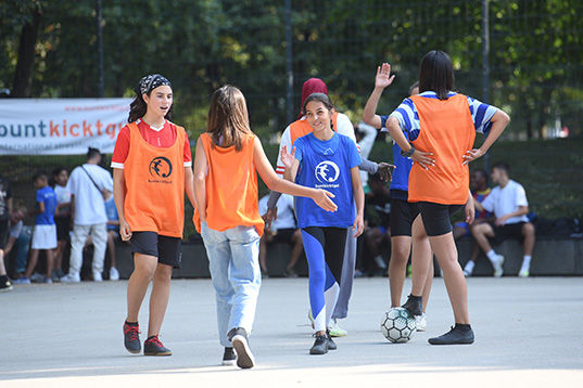 Mädchen in orangenen und blauen Trikots beim Handschlag nach dem Spiel auf einem Hartplatz. Eine von ihnen stellt den Fuß auf einen Fußball.
