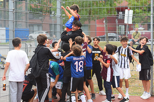 Jungs in Trikots feiern auf einem Sportplatz. In der Mitte eventuell ein Trainer, der einen Jungen hochhebt.