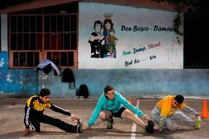 Drei Jungs in Sportkleidung beim Dehnen auf einem Hartplatz. An der Wand dahinter steht "Don Bosco - Damaskus. Run, Jump, Shout. But do not sin." Auf deutsch: Lauf, spring, rufe laut, aber sündige nicht.