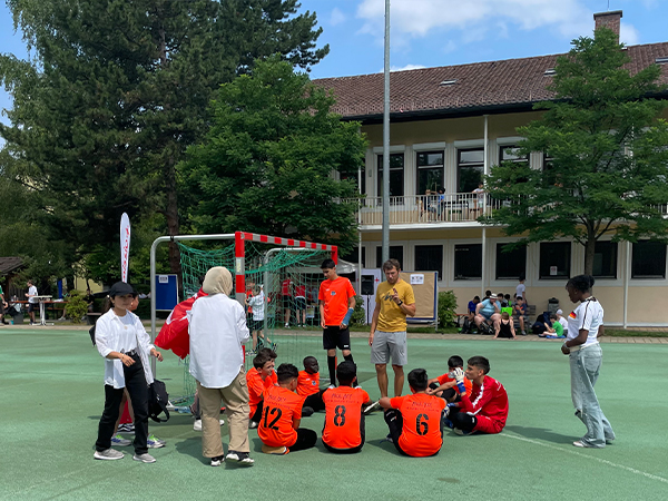 Eine Mannschaft in orangenen Trikots sitzt auf dem Boden, während der Sportlehrer steht und mit der Mannschaft spricht.
