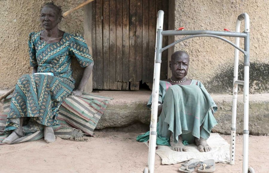 Zwei Frauen sitzen im Lepra-Dorf in Agok auf dem Boden. Eine hat vor sich eine Art Gehkonstruktion stehen.