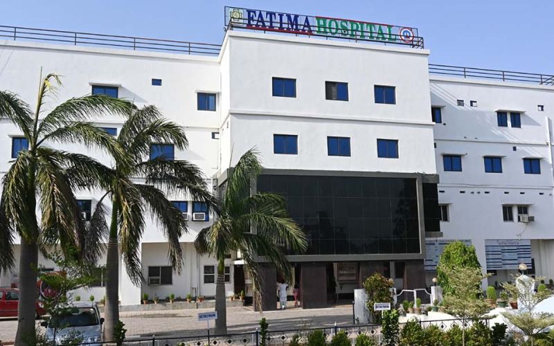 Zu sehen ist das Fatima Krankenhaus, ein großes weißes, rechteckiges Gebäude mit drei großen Palmen im Vordergrund.