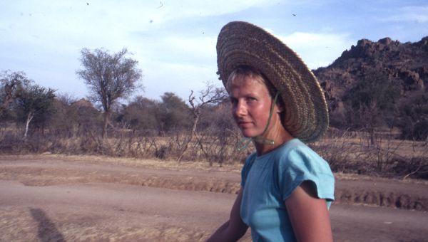 Porträt von Angelika Jakobi 1987 in den Nuba-Bergen: Die junge Frau trägt einen Strohhut, der unter dem Kinn zusammengebunden ist. Ihre Haut ist leicht gebräunt. Sie trägt ein hellblaues T-Shirt.